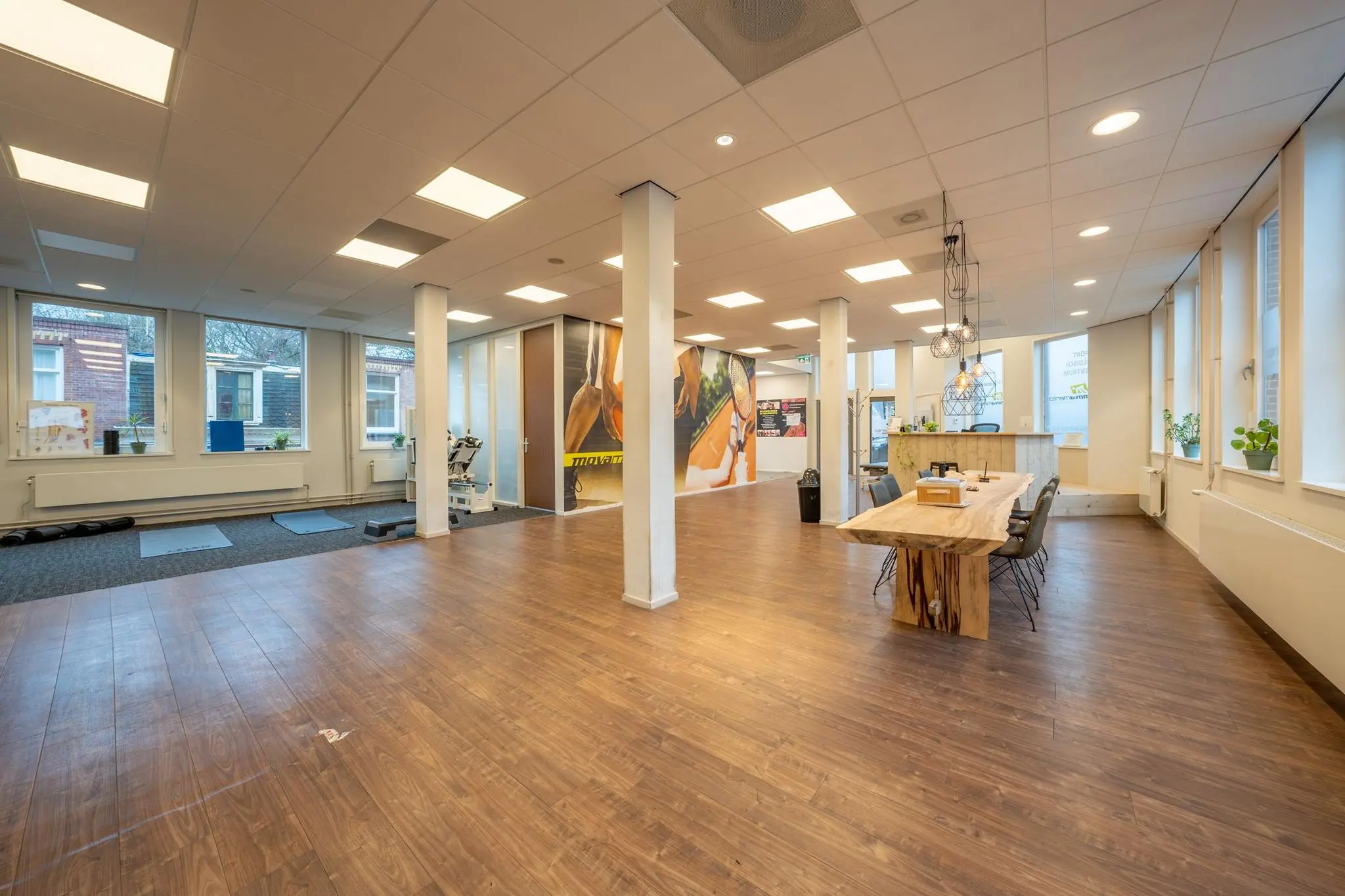Ruim, modern interieur van een praktijkruimte aan de Admiraal de Ruijterweg met houten vloer, fitnessapparatuur en een houten vergadertafel.